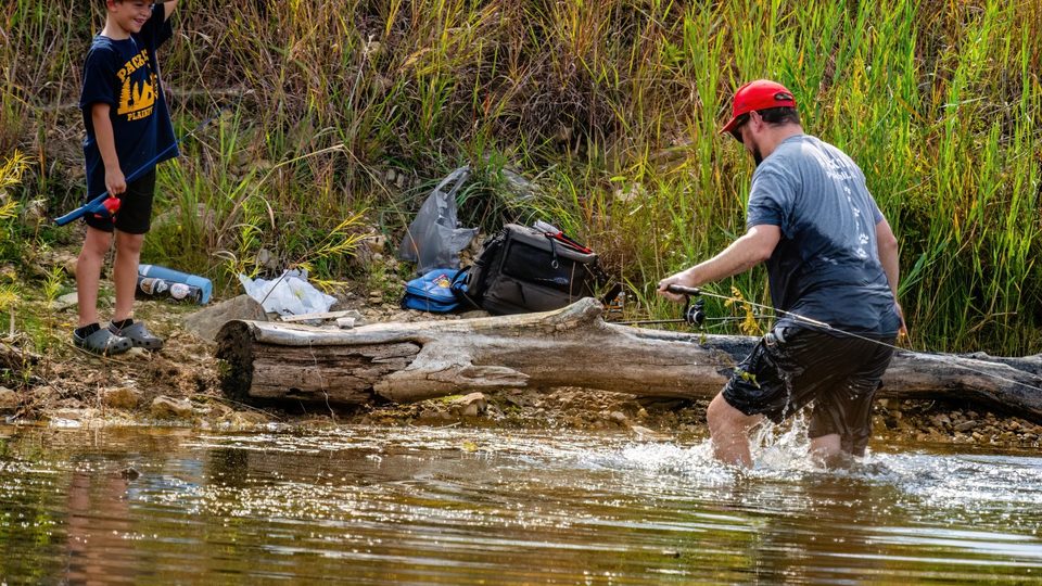 A leader wading into the water to free a fishing line while a laughing Cub Scout watches from the bank