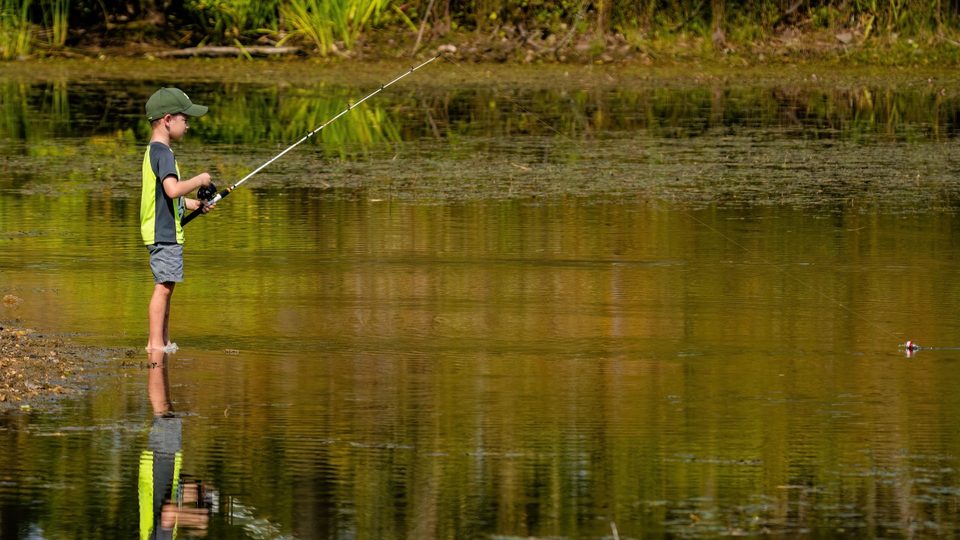 A Cub Scout fishing from the bank, with the rod reflected in the water