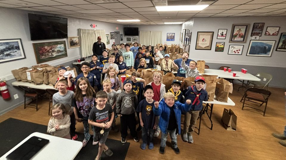 A room full of smiling Cub Scouts behind tables of filled paper grocery bags at the American Legion