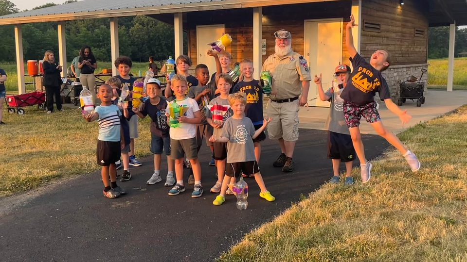 Cub Scouts and a leader at a park pavilion holding homemade bottle rockets, one Scout leaping in the air