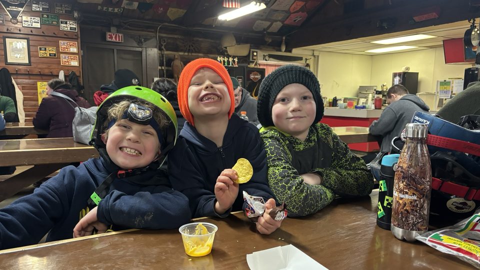 Three grinning Cub Scouts in winter hats sharing chips and cheese at a lodge table