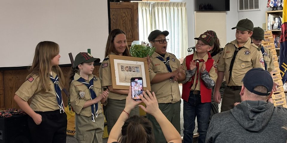 Cub Scouts in uniform cheering as a pack leader holds a framed thank-you and flowers at a pack meeting